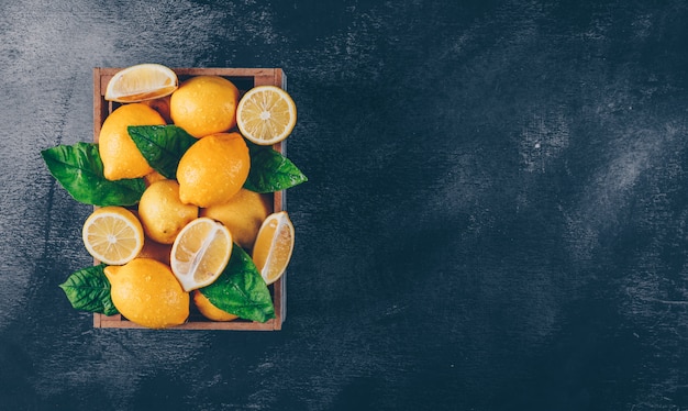 Free photo lemons with slices and leaves in a wooden box on black textured background, top view. copy space for text