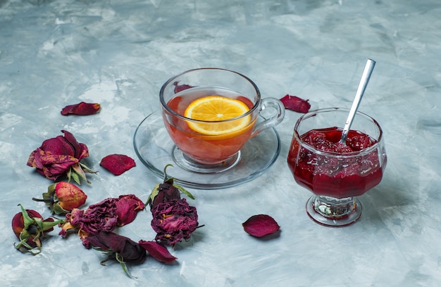 Lemony tea in a cup with dried roses, jam, spoon high angle view on a blue grunge surface