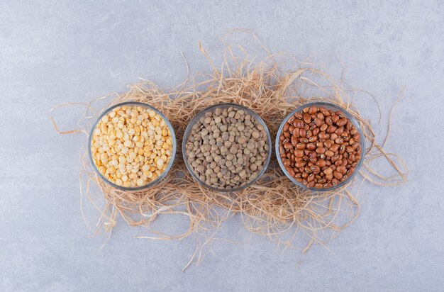 Lentils and red beans portioned into glass bowls on a pile of straw on marble surface