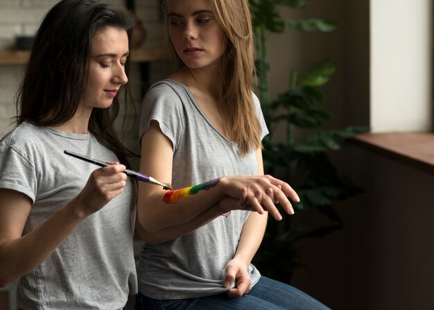 Lesbian woman painting the rainbow flag on her girlfriend's hand with paintbrush