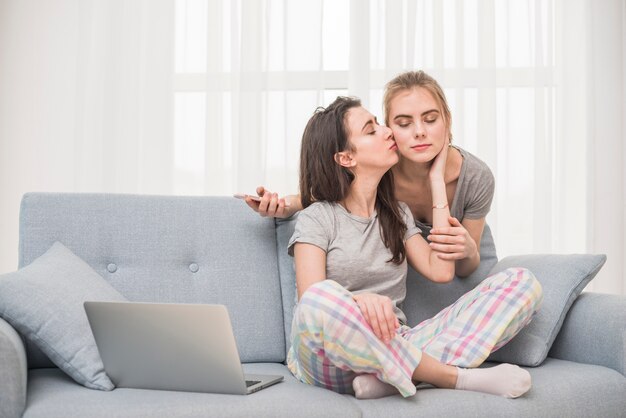 Lesbian woman sitting on sofa kissing her girlfriend holding mobile phone in hand