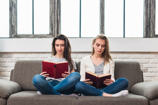 Lesbian young couple sitting on grey sofa with crossed legs reading book