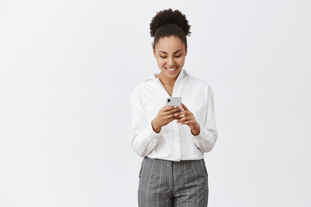 Free photo let me check messages. charming friendly and happy african-american female in white shirt and pants, holding smartphone, gazing entertained and amused at device screen while playing phone game