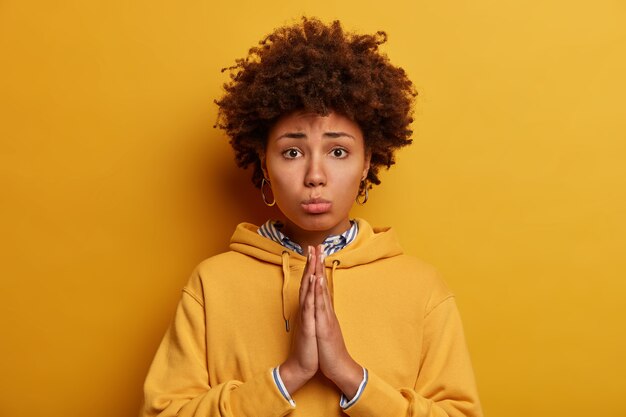 Let me please. Sad pleading African American woman asks for permission, holds hands in pray, says forgive me, poses against yellow wall, wears sweatshirt. Begging and saying forgive me.