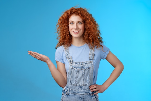 かっこいい商品をお見せしましょう。魅力的な陽気な自信を持って赤毛の女性モデルの巻き毛の髪型現在の顧客オブジェクトは、立っている青い背景を宣伝する手のひらの空白の青いコピースペースを保持します。