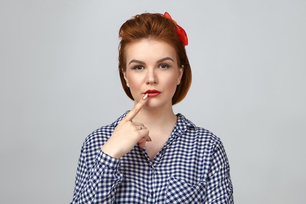 Let me think. Isolated picture of fashionable elegant young European lady with ginger hair having deep in thoughts pensive facial expression, touching lips, trying to remember something important