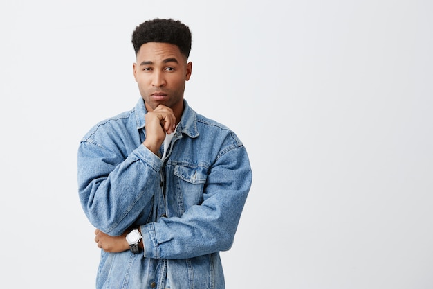 Let me think. Isolated portrait of young attractive tan-skinned man with afro hairstyle in denim jacket holding chin with hand, looking in camera with thoughtful face expression. Copy space