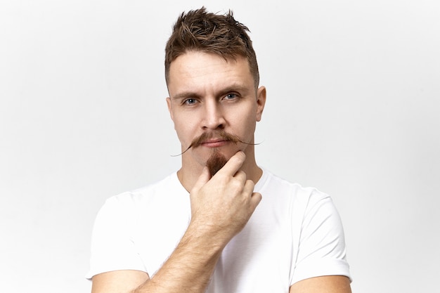 Let me think. Pensive thoughtful young Caucasian male with stylish mustache touching his goatee beard while pondering, having deep in thoughts look. Body language and human facial expressions