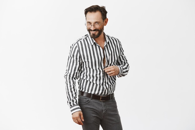 Free photo let us continue conversation in bedroom honey portrait of flirty handsome adult man with beard unbuttoning shirt and gazing sensually at camera hinting and foreplaying with mistress over grey wall