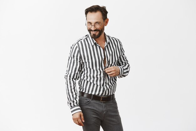 Let us continue conversation in bedroom honey Portrait of flirty handsome adult man with beard unbuttoning shirt and gazing sensually at camera hinting and foreplaying with mistress over grey wall