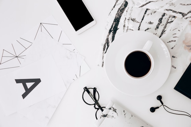 Letter a; eyeglasses; smartphone; earphone; coffee cup on white desk