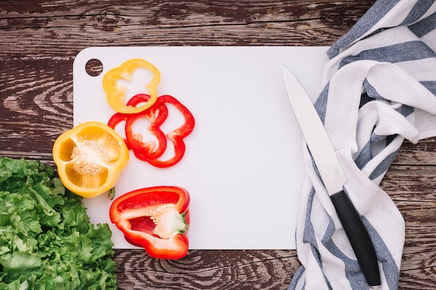 Lettuce and bell peppers on white chopping board with knife and napkin over the wooden desk