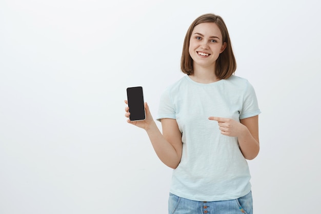 Life changed after this phone. Portrait of friendly-looking delighted charming woman with short brown hair in casual light t-shirt showing cellphone screen and pointing at smartphone smiling