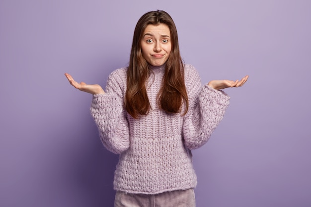 Free photo life perception concept. unaware european female shrugs shoulders, makes decision, wears violet sweater, has hesitant facial expression, stands and gestures in , isolated over purple wall