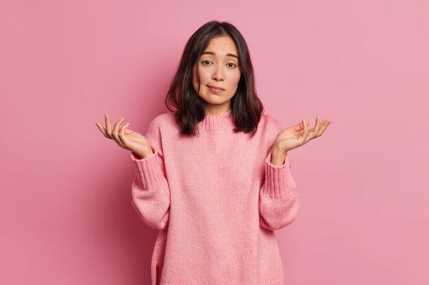 Life perception and hard decision concept. Puzzled clueless young Asian woman spreads palms and poses uncertian indoor expresses doubt wears knitted sweater 
