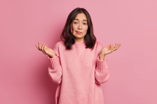 Life perception and hard decision concept. Puzzled clueless young Asian woman spreads palms and poses uncertian indoor expresses doubt wears knitted sweater 