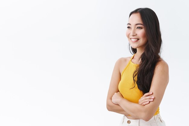 Lifestyle beauty and emotions concept Cheerful selfassured young eastasian woman in yellow top standing halfturned looking sideways and giggle laughing joyfully smiling feeling happy