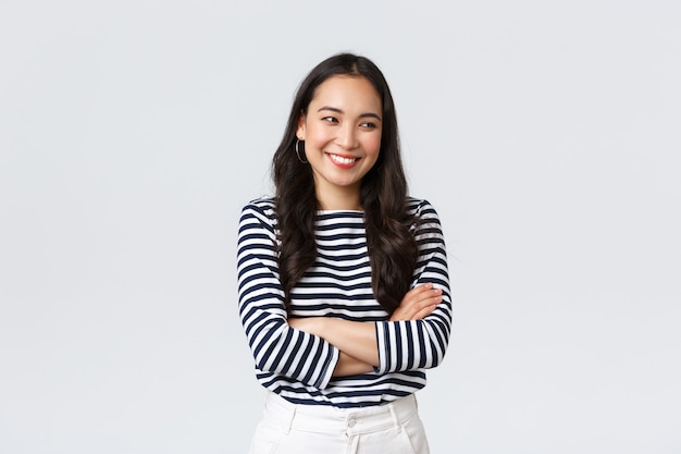 Lifestyle, beauty and fashion, people emotions concept. Cheerful cute and shy asian girl cross arms chest modest pose, looking away as laughing and smiling silly, stand white background