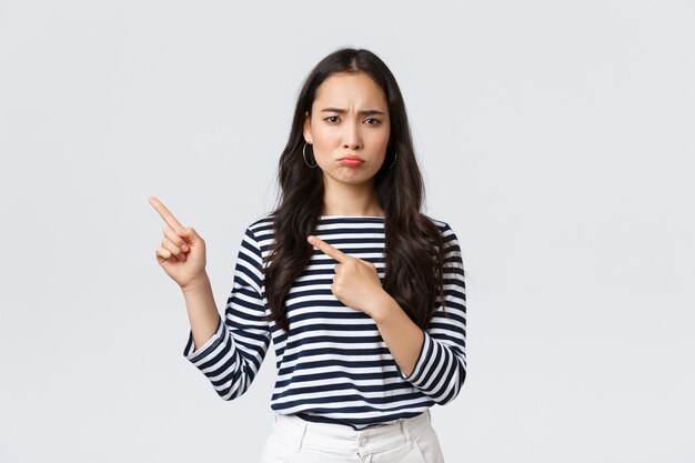 Lifestyle, beauty and fashion, people emotions concept. Disappointed sad asian girl complaining, feeling regret or jealousy as pouting uneasy and pointing fingers left at promo banner