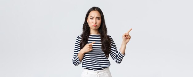 Lifestyle beauty and fashion people emotions concept Disappointed upset young asian woman pouting and frowning displeased pointing fingers right top as complaining about product or service