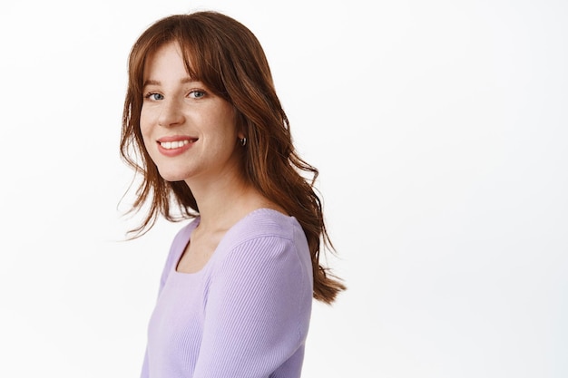 Lifestyle and beauty. Happy young woman with freckles and white smile, standing in profile, turn face at camera with carefree face, standing in blouse against white background