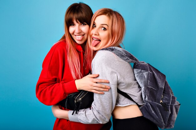 Lifestyle bright portrait of happy couple of hipster girls, showing tongues and hug each other, best friends having fun, blue wall, wearing hoodies and backpack.
