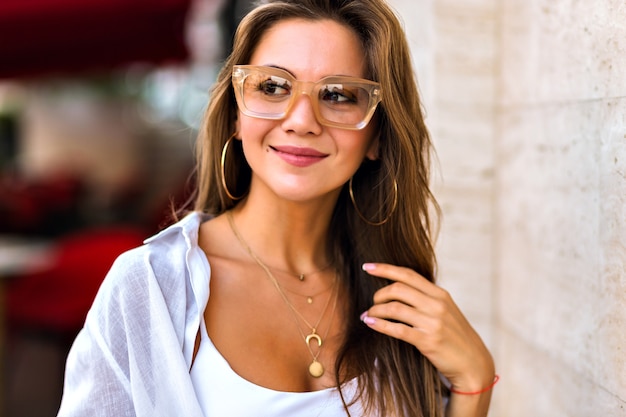 Lifestyle city portrait of amazing attractive young brunette woman wearing beige trendy clear glasses and gold jewelry, soft warm colors, minimalism style.