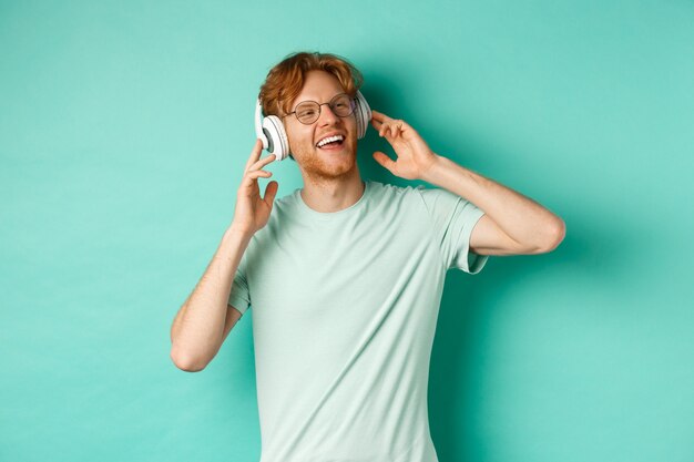 Lifestyle concept. Happy young man with ginger hair dancing and having fun, listening music on wireless headphones and smiling pleased, turquoise background.