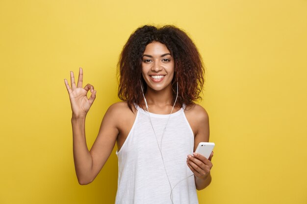 Концепция образа жизни - портрет красивой афро-американской женщины радостно слушать музыку на мобильном телефоне. Желтый пастельный фон студии. Копирование пространства.