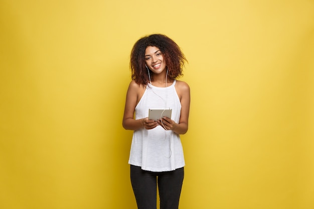 Lifestyle Concept - Portrait of beautiful African American woman joyful listening to music on tablet. Yellow pastel studio background. Copy Space.