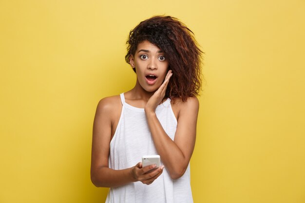 라이프 스타일 개념-휴대 전화에 뭔가 충격적인 아름 다운 아프리카 계 미국인 여자의 초상화. 노란 파스텔 스튜디오 배경입니다. 공간을 복사하십시오.