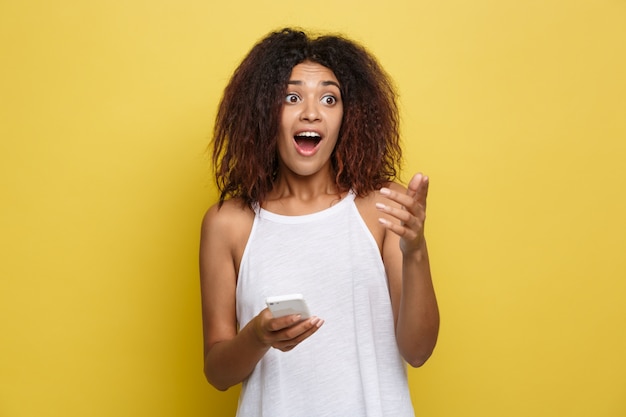 Lifestyle Concept - Portrait of beautiful African American woman shocking with something on mobile phone. Yellow pastel studio background. Copy Space.