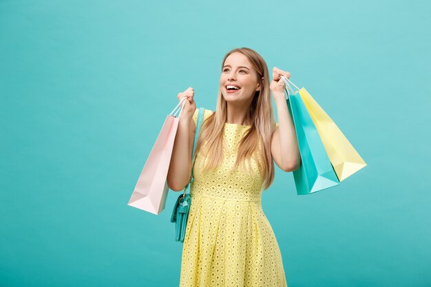 ライフスタイルの概念：買い物袋でポーズをとって、青い背景の上にカメラを見て黄色の夏のドレスでショックを受けた若い魅力的な女性の肖像画。