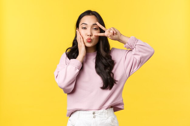 Lifestyle, emotions and advertisement concept. Coquettish and cute, stylish asian woman showing kawaii peace sign over eye and folding lips for kiss, feeling beautiful, yellow background.