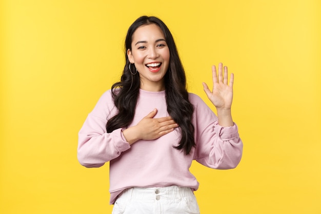 Lifestyle, emotions and advertisement concept. Honest and sincere happy smiling asian woman promise tell truth, swear on her heart and raising one hand, standing yellow background.