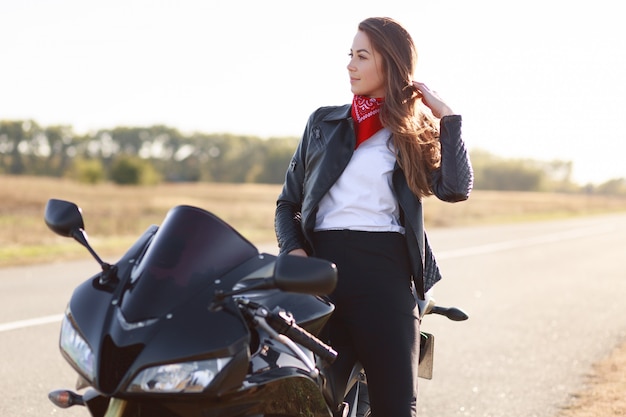 Free photo lifestyle, extreme and people concept. sideways shot of pretty thoughtful young female driver dressed in fashionable clothes, stands near favourite motorbike, poses outdoor, enjoys fast driving