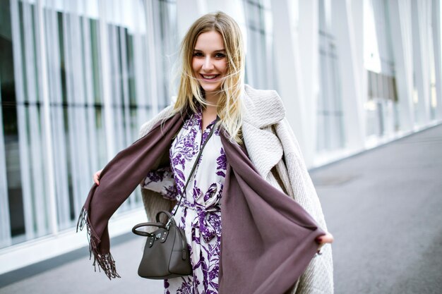 Lifestyle fashion portrait of pretty elegant woman wearing trendy stylish outfit, violet long scarf, cashmere luxury coat and midi dress, smiling end enjoying, stay in front of modern business center