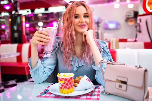 Lifestyle indoor image of stylish young pretty woman with wavy unusual pink hairs and natural make up, wearing cute blue dress and denim jacket, enjoy her tasty American dinner.