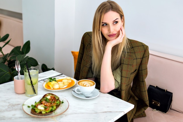 Ritratto dell'interno di stile di vita di elegante giovane donna bionda di affari che gode del suo brunch al caffè hipster, chiedendo cameriere e sorridente