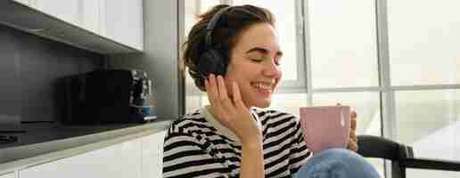 Free photo lifestyle and music concept happy smiling woman with cup of tea sits in kitchen and touches