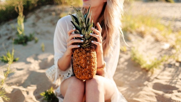Изображение жизни на открытом воздухе смеясь над красивой женщиной с сочным ананасом, расслабляющимся на солнечном пляже. Модный летний наряд