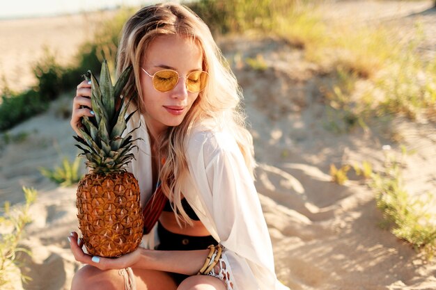 햇볕이 잘 드는 해변에서 편안 하 게 육즙 파인애플과 웃는 예쁜 여자의 라이프 스타일 야외 사진. 트렌디 한 여름 의상