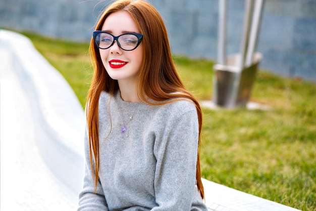 Free photo lifestyle outdoor portrait of pretty ginger girl wearing clear glass and cashmere grey sweater, posing at the park, autumn winter style.