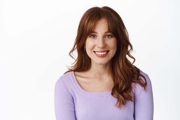 Lifestyle and people concept. Smiling beautiful young woman in stylish clothes, with freckles and bangs, white teeth and natural face expression, standing against white background.