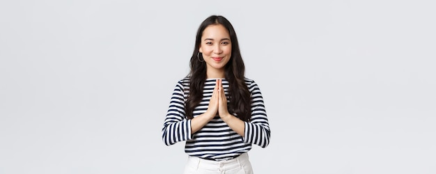 Lifestyle people emotions and casual concept Cute asian girl in casual outfit smiling as saying namaste holding hands in pray pleading or greeting guests with polite bow
