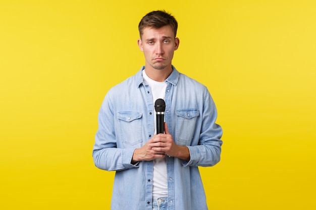 Lifestyle, people emotions and summer leisure concept. Gloomy sad blond man, student with microphone looking upset, singing heartbreaking song, standing yellow background.