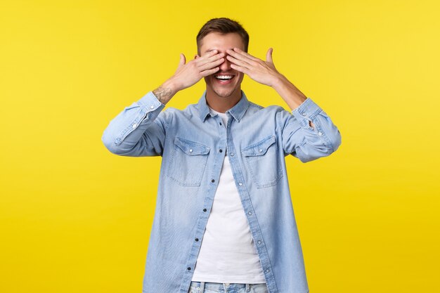 Lifestyle, people emotions and summer leisure concept. Smiling happy handsome man shut eyes with hands and anticipating for surprise, standing upbeat at birthday party, yellow background.