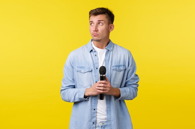 Lifestyle, people emotions and summer leisure concept. Thoughtful dreamy handsome man looking away, thinking as holding microphone, standing yellow background indecisive