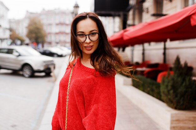 Lifestyle portrait of brunette woman with long hairs in red cardigan walking through street. Windy hairs. Stylish eyeglasses.