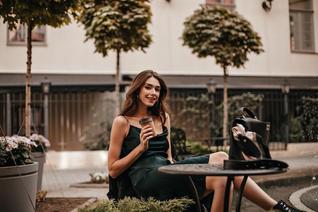 Free photo lifestyle portrait of charming young brunette in green silk dress sitting at city cafe terrace and drinking morning coffee light building and trees background